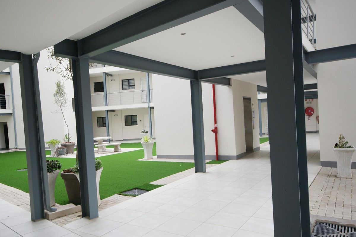 A well-maintained apartment complex courtyard featuring a covered walkway with steel structural supports, tiled flooring, and a neat artificial lawn area.
