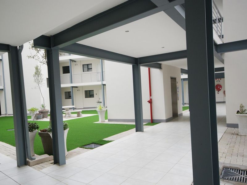 A well-maintained apartment complex courtyard featuring a covered walkway with steel structural supports, tiled flooring, and a neat artificial lawn area.
