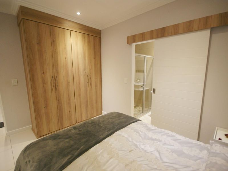 A modern bedroom featuring built-in wood-finish cupboards, sleek tiled flooring, and a sliding door providing access to an en-suite bathroom.