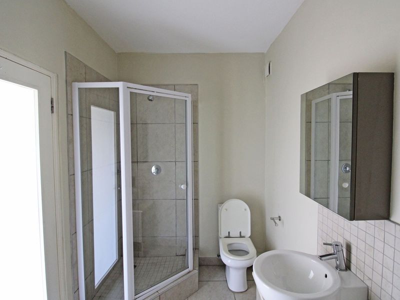 A functional bathroom featuring a corner shower enclosure, a wall-mounted basin with a mirrored cabinet, and a toilet, finished with neutral floor tiles.