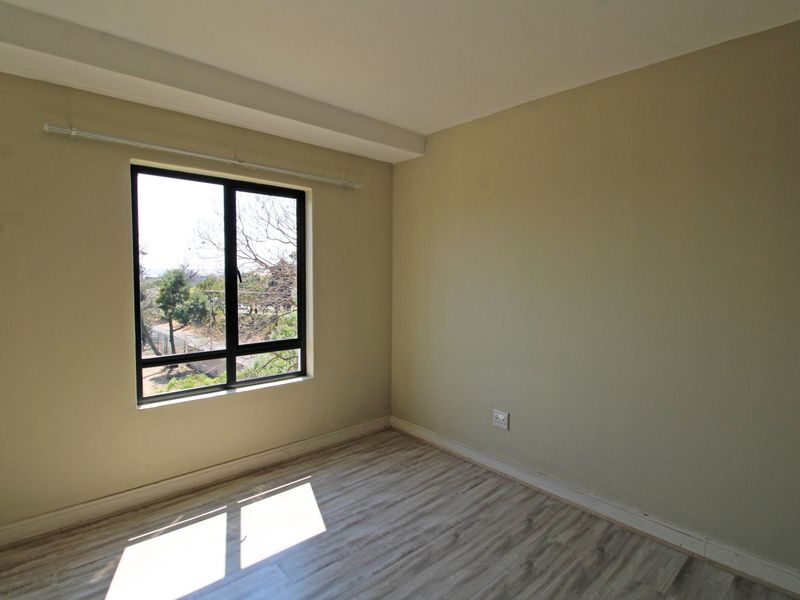 A bright, empty bedroom featuring light-toned laminate flooring, neutral wall paint, and an aluminium-framed window with a curtain rod.