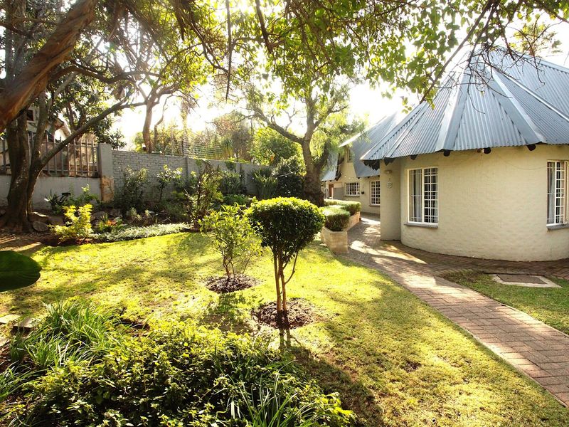 A well-maintained rear garden featuring a brick paved pathway, mature trees, and a house with a distinctive corrugated metal roof and casement windows.