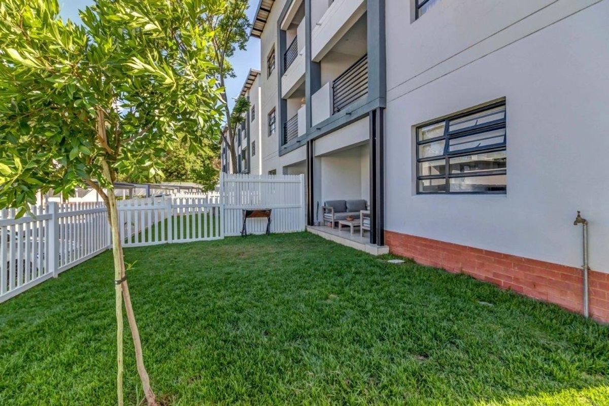 A private rear garden area featuring a lawn, white picket fencing, and a covered patio space attached to the apartment building.