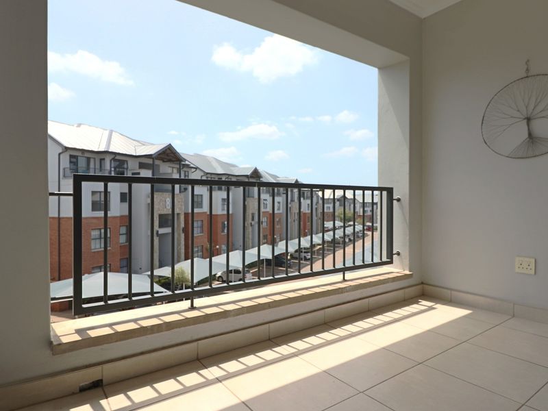 A clean, covered balcony featuring tiled flooring and a secure metal balustrade, offering an open view of the apartment complex grounds.