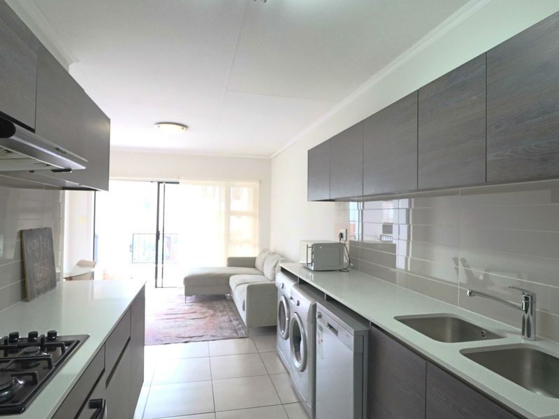 Modern open-plan kitchen featuring dark wood cabinetry, stone countertops, gas hob, and integrated laundry appliances, flowing into a bright living area with balcony access.
