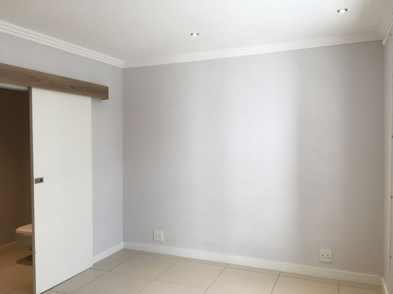 A clean, empty bedroom featuring neutral tiled flooring, recessed downlighting, and a modern sliding door leading to an en-suite bathroom.