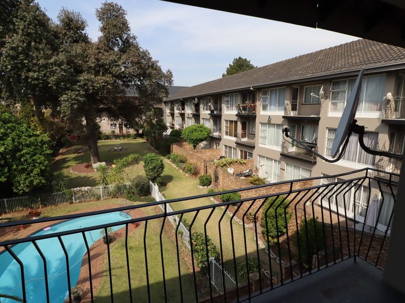 A balcony view overlooking the apartment complex grounds, featuring a swimming pool, landscaped garden, and mature trees.