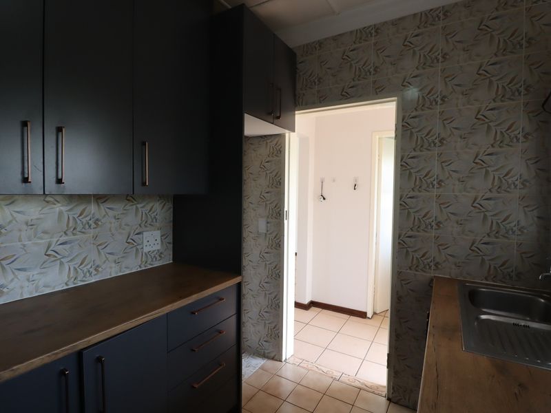 A compact kitchen featuring dark cabinetry, wooden countertops, a stainless steel sink, and patterned tiled backsplash, leading into an adjacent hallway.
