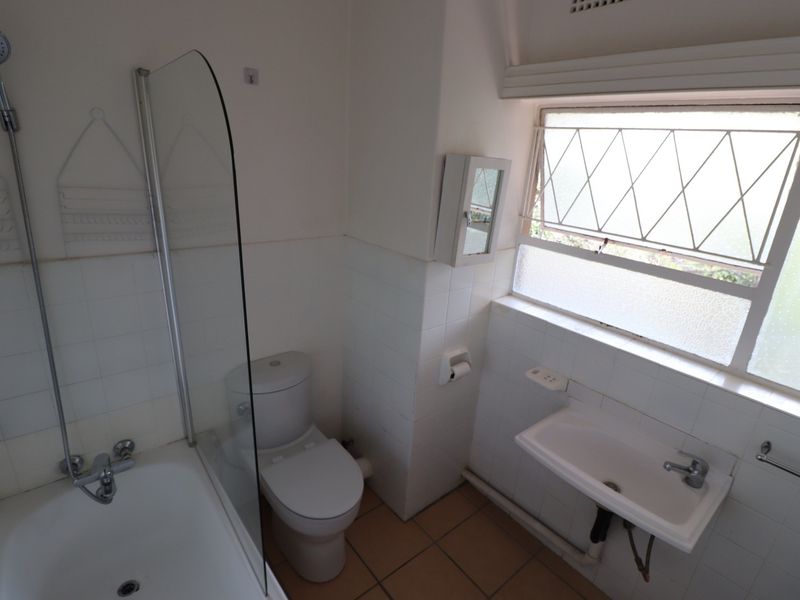 A functional bathroom featuring a bathtub with shower attachment, glass screen, toilet, and wall-mounted basin with tiled finishes.