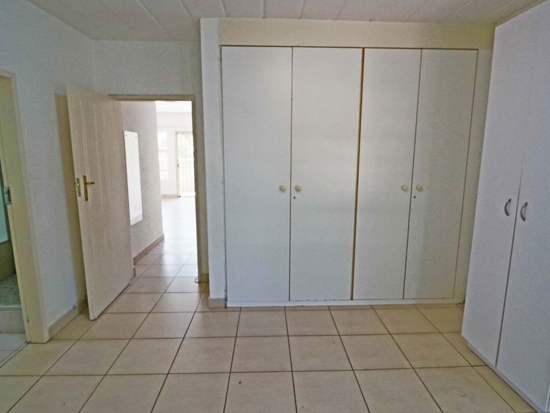 A spacious, empty bedroom featuring neutral tiled flooring and extensive built-in cupboard storage, with direct access to an adjoining bathroom and hallway.
