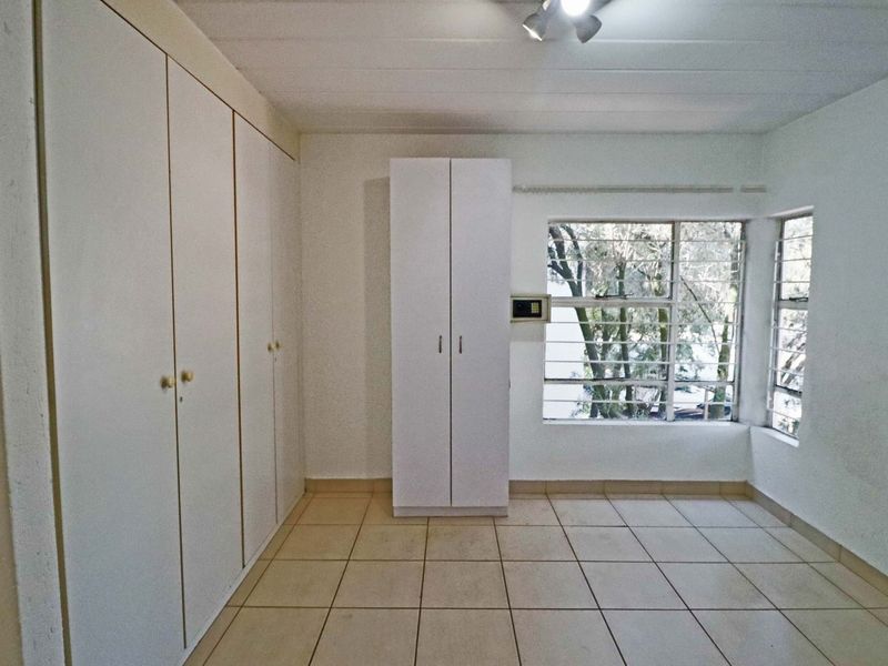 A bright, empty bedroom featuring tiled flooring, extensive built-in cupboard storage, a wall-mounted safe, and windows fitted with security bars.