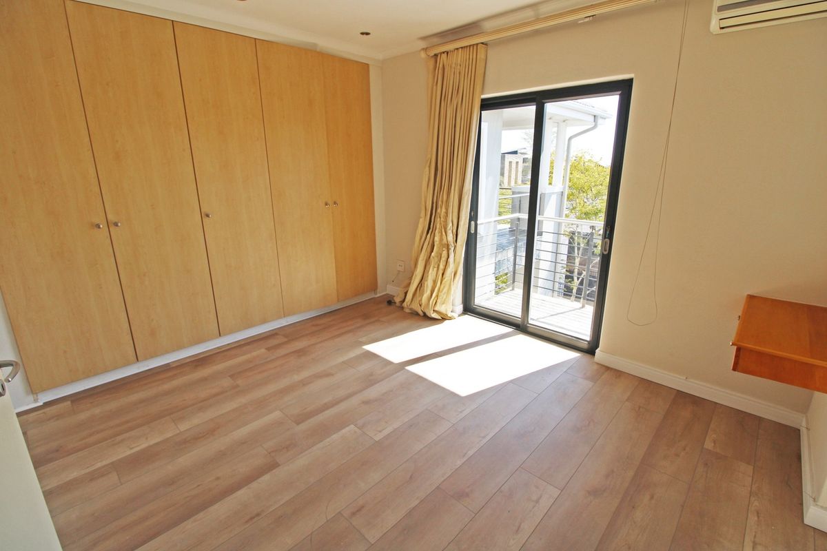 A bright, empty bedroom featuring light wood-look laminate flooring, extensive built-in cupboards, and a sliding door providing access to a private balcony.