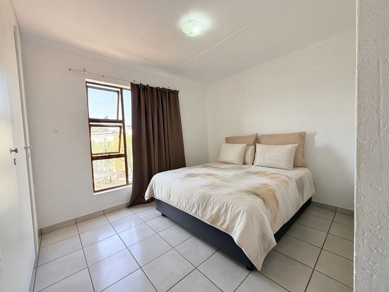 A simple bedroom featuring tiled flooring, a built-in cupboard, and a window providing natural light.