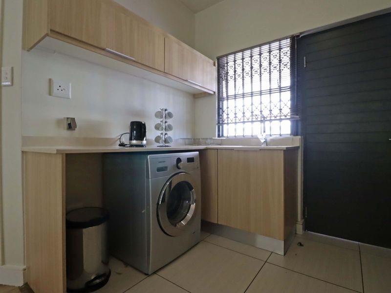 A functional laundry area featuring light wood-finish cabinetry, a laminate countertop, tiled flooring, and a secure window with a metal security gate.