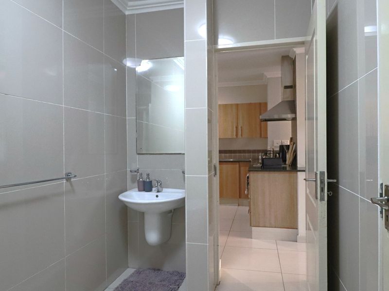 A clean, tiled bathroom featuring a pedestal basin and wall-mounted mirror, with a view through to the adjacent kitchen area.