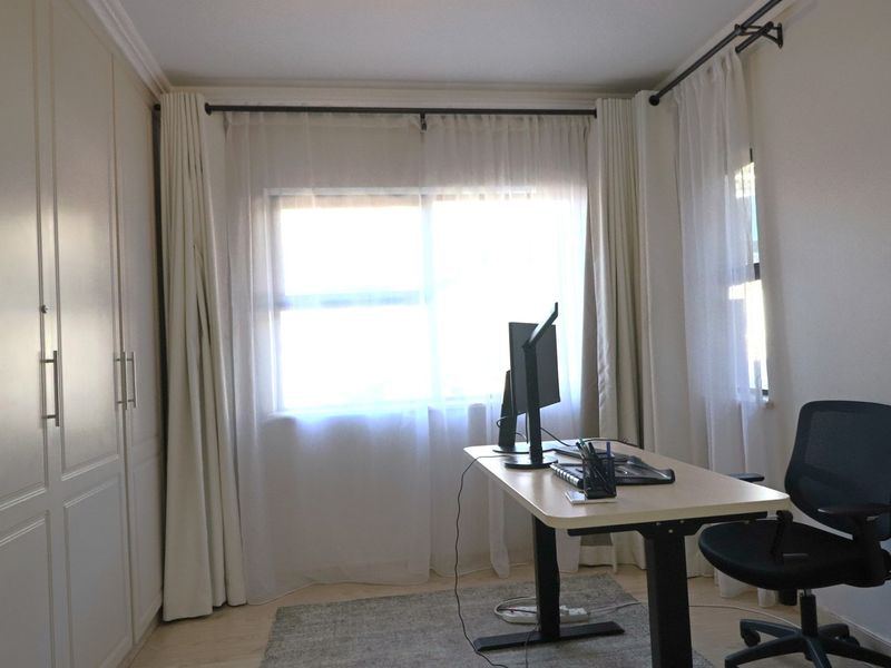 A functional bedroom featuring built-in cupboards, laminate flooring, and large windows with curtain rods, currently utilized as a home office space.