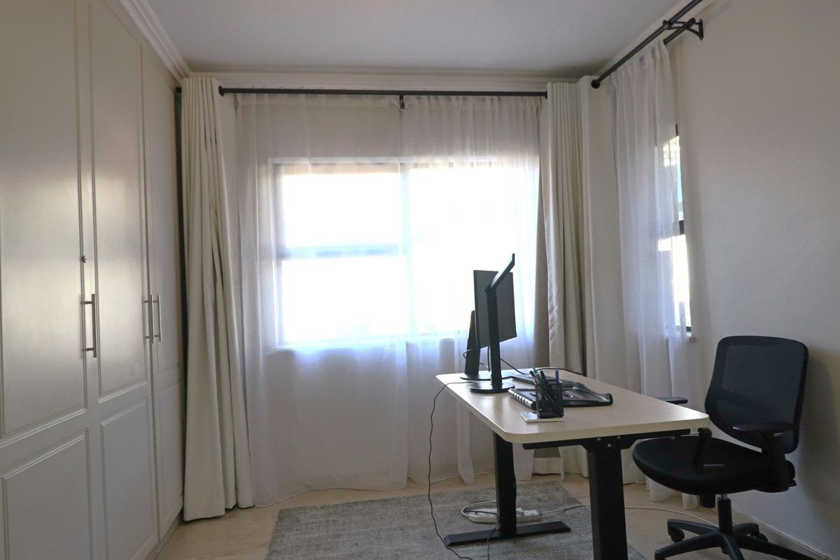 A functional bedroom featuring built-in cupboards, laminate flooring, and large windows with curtain rods, currently utilized as a home office space.