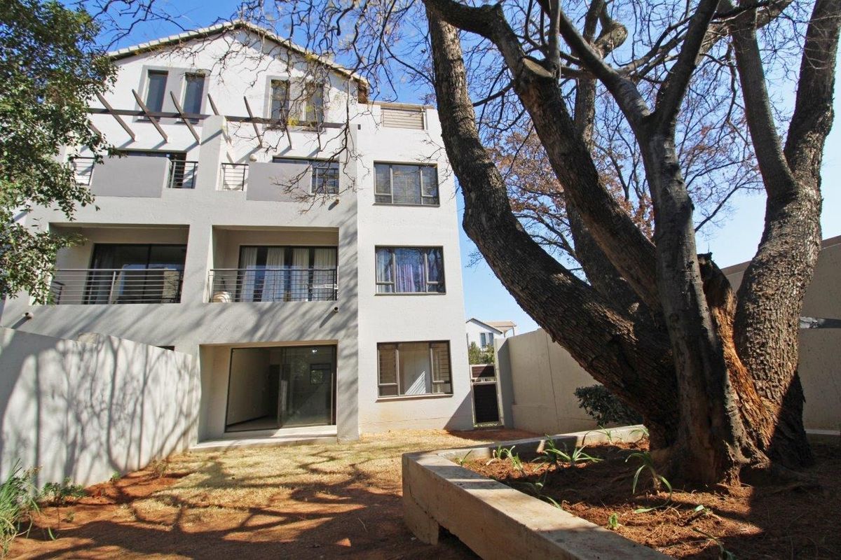 Rear view of a multi-story apartment building featuring balconies, sliding glass doors, and a landscaped garden area with a large mature tree.