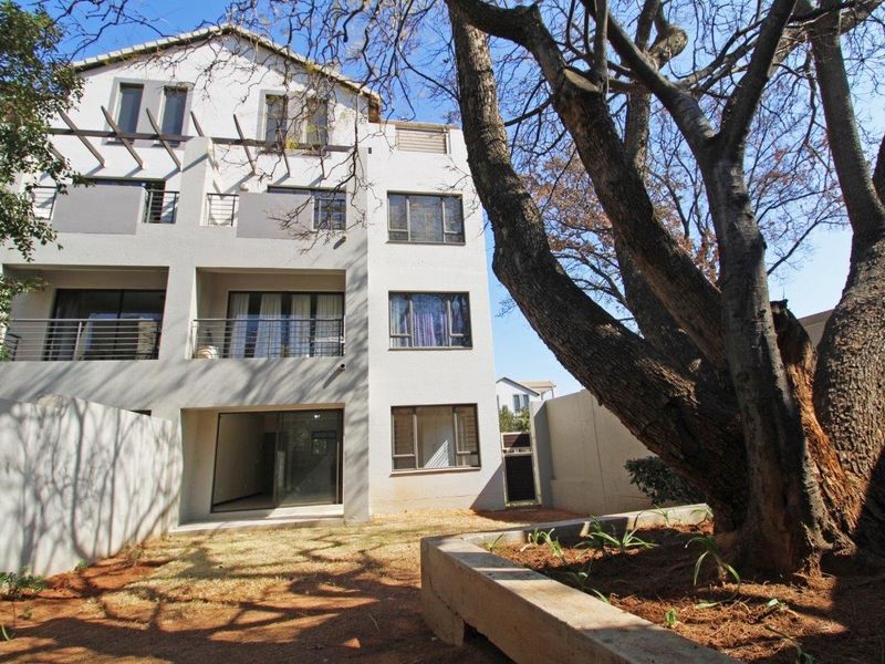 Rear view of a multi-story apartment building featuring balconies, sliding glass doors, and a landscaped garden area with a large mature tree.