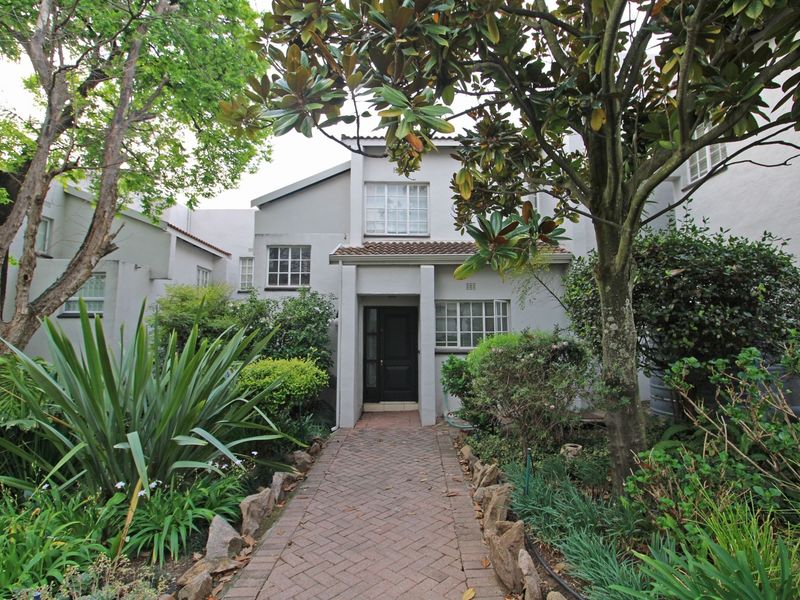Front elevation of a townhouse featuring a brick paved walkway leading to the entrance, surrounded by established garden landscaping and mature trees.