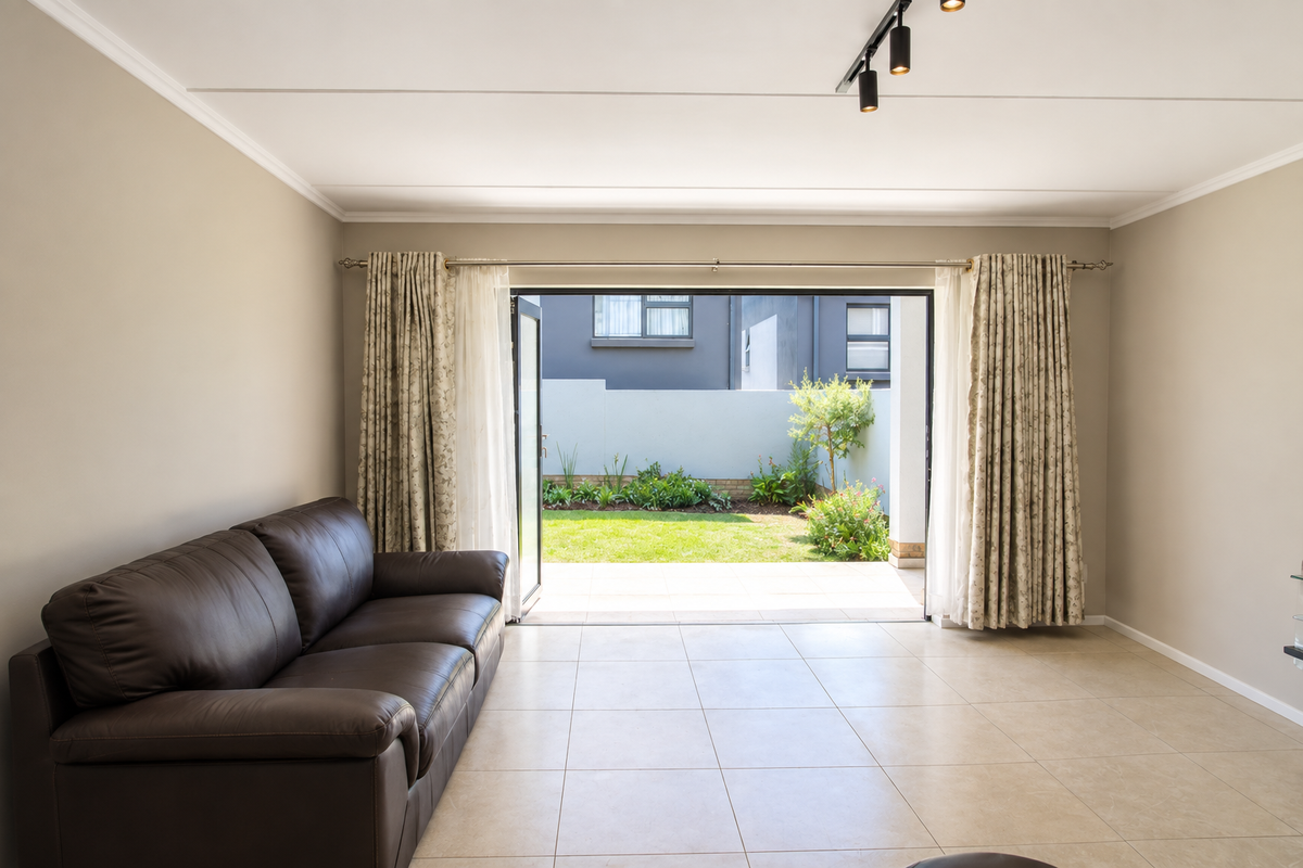 A bright living room featuring neutral tiled flooring, modern track lighting, and large sliding glass doors that open onto a private garden patio.