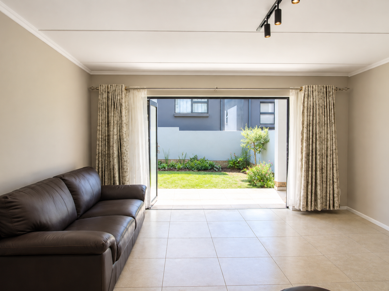 A bright living room featuring neutral tiled flooring, modern track lighting, and large sliding glass doors that open onto a private garden patio.