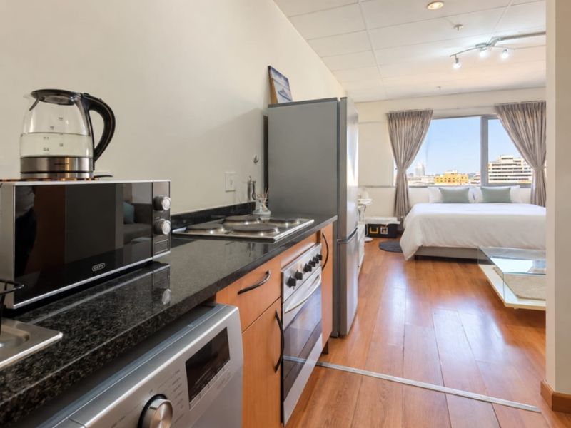 Open-plan studio apartment featuring a compact kitchen with granite countertops and integrated appliances, flowing into a bedroom area with large windows and city views.