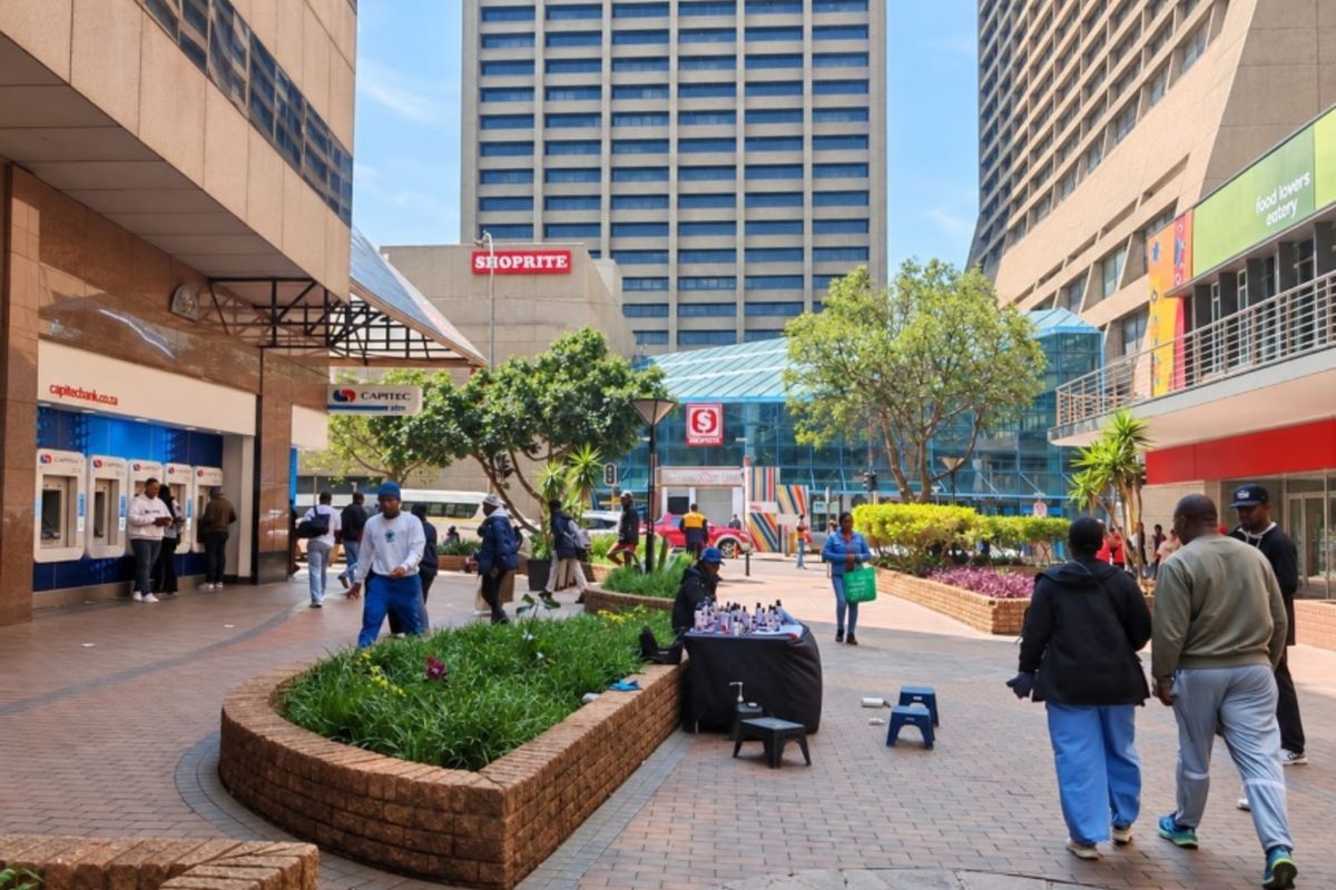 A bustling urban pedestrian plaza featuring paved walkways, brick landscaping planters, and access to various commercial storefronts and banking facilities.