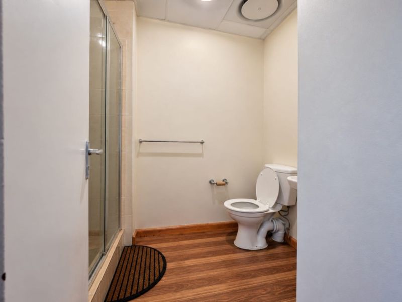A functional bathroom featuring a shower enclosure, toilet, and laminate flooring, with a drop ceiling and neutral wall finishes.