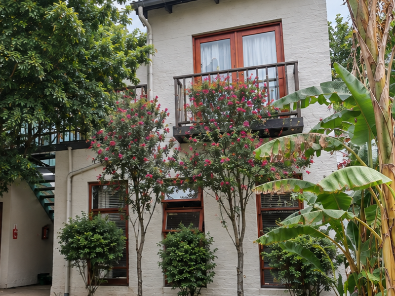 A two-story residential facade featuring timber-framed windows and doors, a balcony with metal railings, and an external staircase set within a landscaped garden.