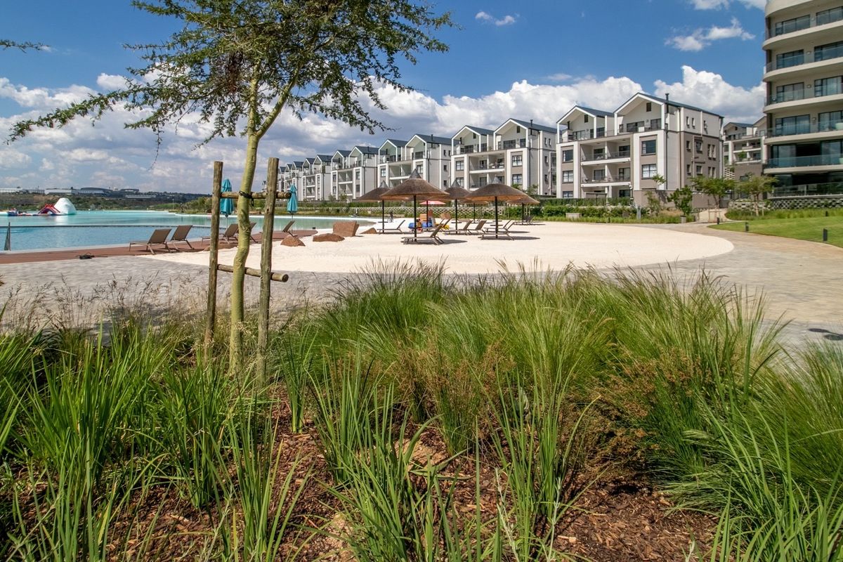 A well-maintained communal pool area featuring a beach-style entry, landscaped gardens, and paved walkways within a modern apartment complex.