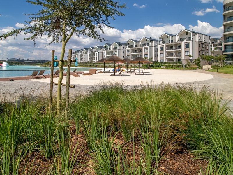 A well-maintained communal pool area featuring a beach-style entry, landscaped gardens, and paved walkways within a modern apartment complex.