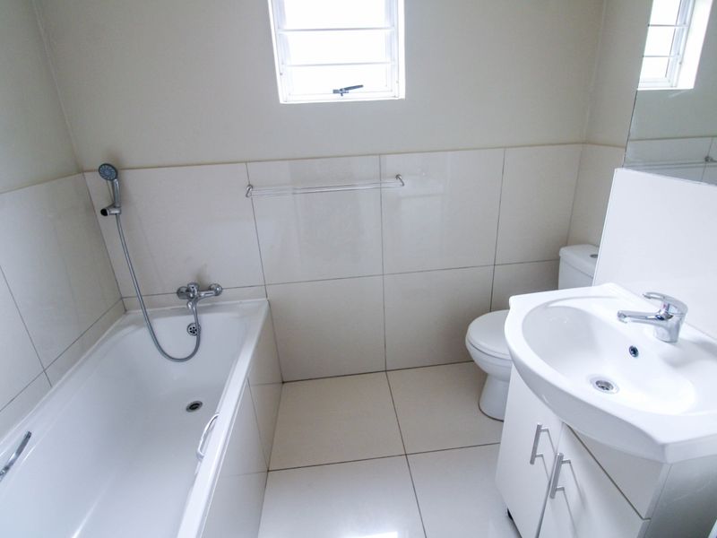 A clean, neutral-toned bathroom featuring a bathtub with handheld shower, a vanity unit with integrated basin, and tiled finishes throughout.