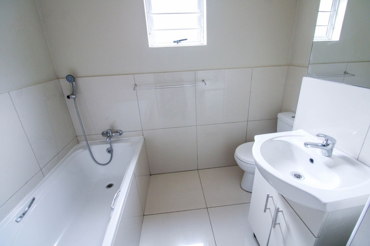 A clean, neutral-toned bathroom featuring a bathtub with handheld shower, a vanity unit with integrated basin, and tiled finishes throughout.