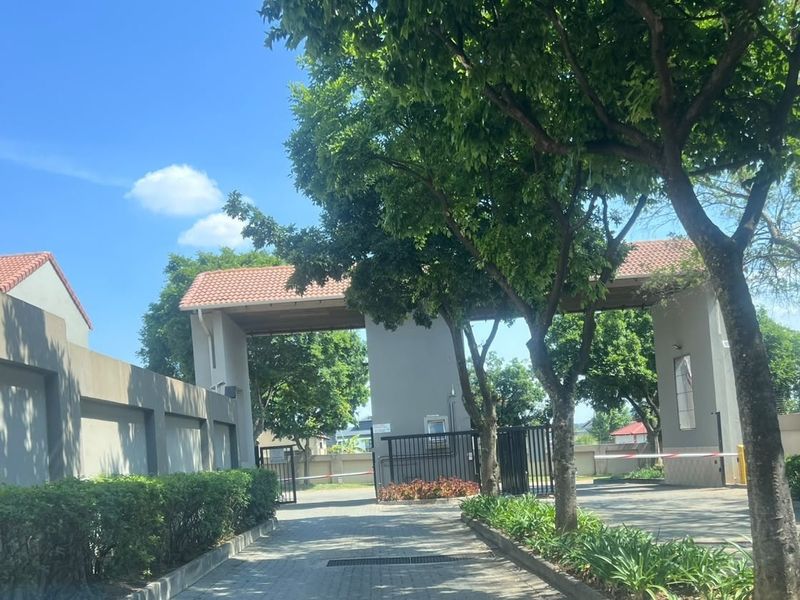 A view of the secure gated entrance to the townhouse complex, featuring a paved driveway, perimeter walls, and established landscaping.