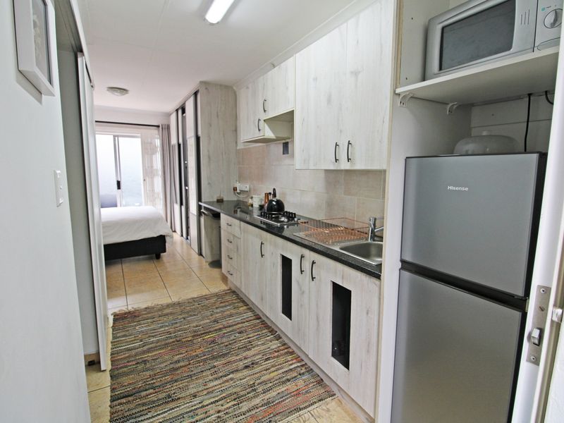 Compact open-plan kitchen featuring light wood-look cabinetry, stone countertops, and integrated appliances, flowing into a bedroom area with sliding door access.