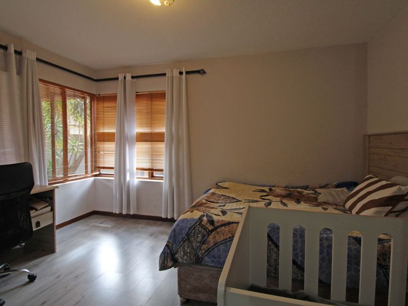A bedroom featuring light-toned laminate flooring, corner windows with wooden venetian blinds, and neutral wall finishes.