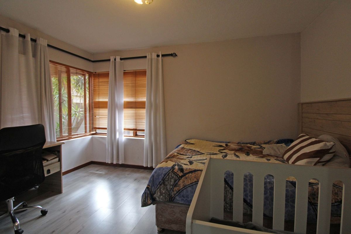 A bedroom featuring light-toned laminate flooring, corner windows with wooden venetian blinds, and neutral wall finishes.