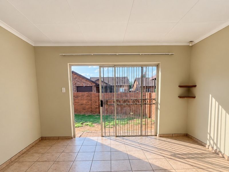 A bright, empty living room featuring tiled flooring, built-in wall shelving, and a sliding glass door with a security gate leading to the rear garden.