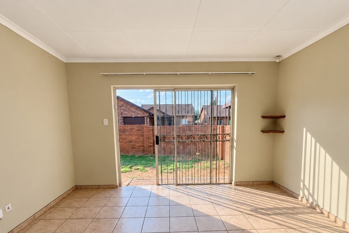 A bright, empty living room featuring tiled flooring, built-in wall shelving, and a sliding glass door with a security gate leading to the rear garden.