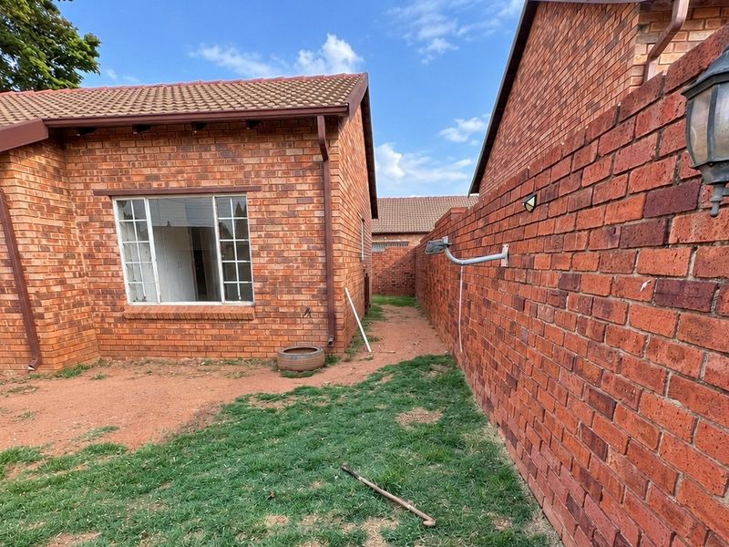 Rear exterior view of a face brick townhouse featuring a small garden area, security gate, and perimeter wall with mounted lighting.