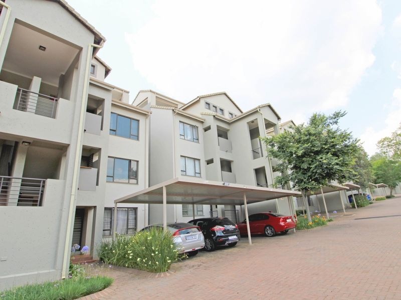 Exterior view of a multi-story apartment complex featuring covered parking bays, paved driveway, and private balconies with aluminium window frames.