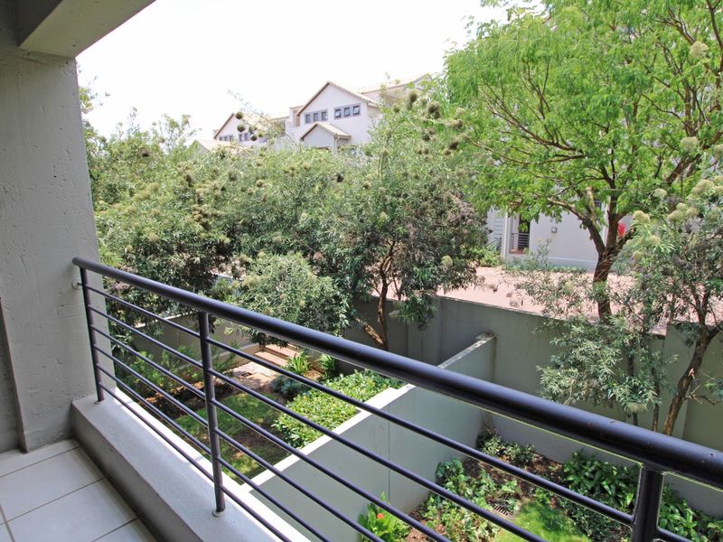 A private balcony featuring a metal balustrade and tiled flooring, overlooking a landscaped garden area and neighboring residential complex.