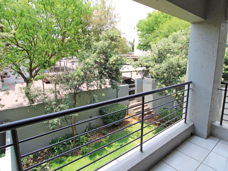 A covered balcony featuring tiled flooring and a metal balustrade, overlooking a landscaped garden area and complex parking facilities.