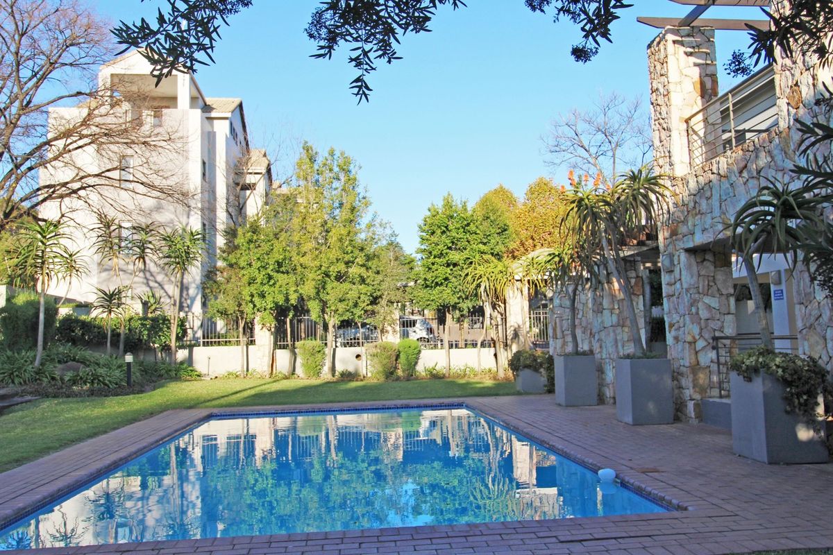 A well-maintained communal swimming pool area featuring stone-clad pillars, brick paving, and established garden landscaping within an apartment complex.