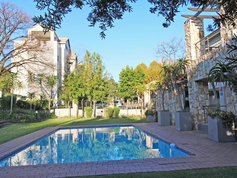 A well-maintained communal swimming pool area featuring stone-clad pillars, brick paving, and established garden landscaping within an apartment complex.