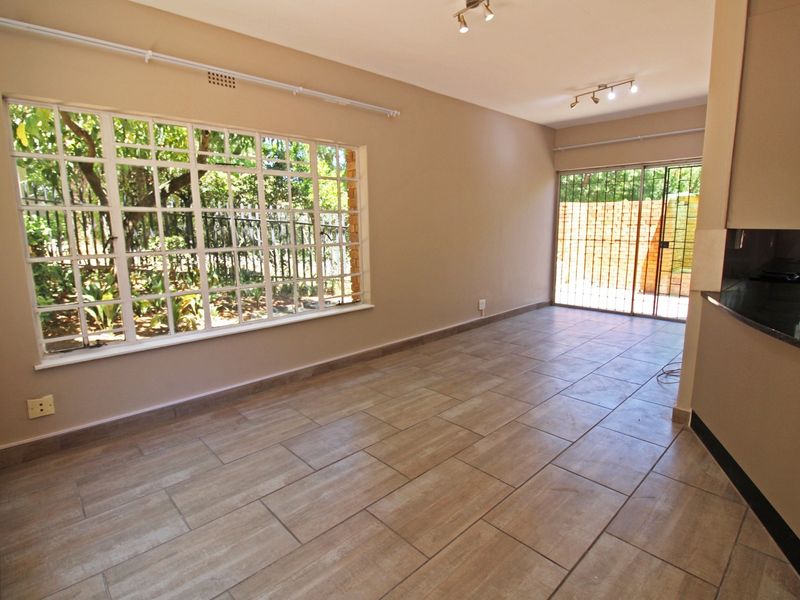 Bright open-plan living area featuring modern tiled flooring, large windows, and a sliding door leading to an outdoor space, adjacent to a kitchen area.