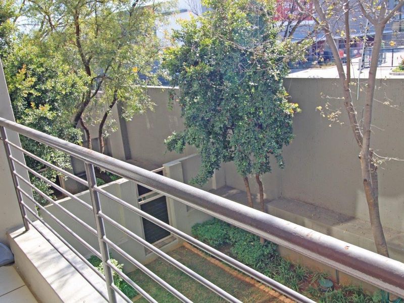 A private balcony featuring a metal balustrade and tiled flooring, overlooking a landscaped garden area with mature trees and boundary walls.