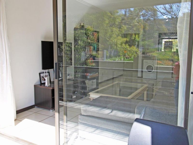 A view through a sliding glass door onto a balcony with tiled flooring and metal railings, overlooking a garden area.