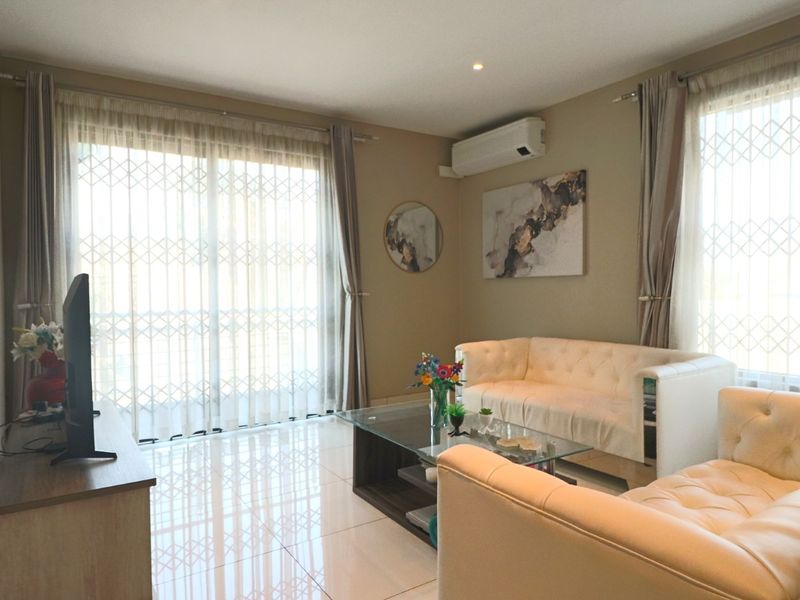 A well-maintained living room featuring tiled flooring, recessed lighting, and a wall-mounted split-system air conditioner, with large windows fitted with security gates.